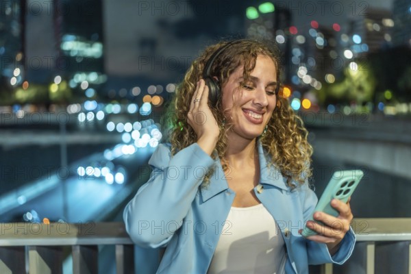 Woman with headphones and smartphone enjoying music while smiling cheerfully, standing on a bridge with city lights blurring in the background at night
