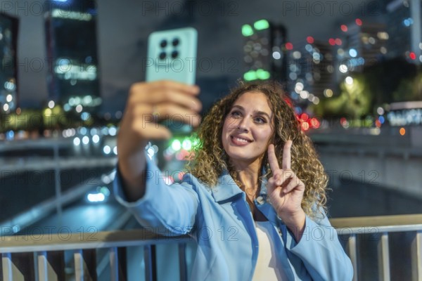 Young woman with curly hair smiling and flashing a peace sign while taking a nighttime city selfie with her smartphone, bokeh lights blurring the urban background and mood