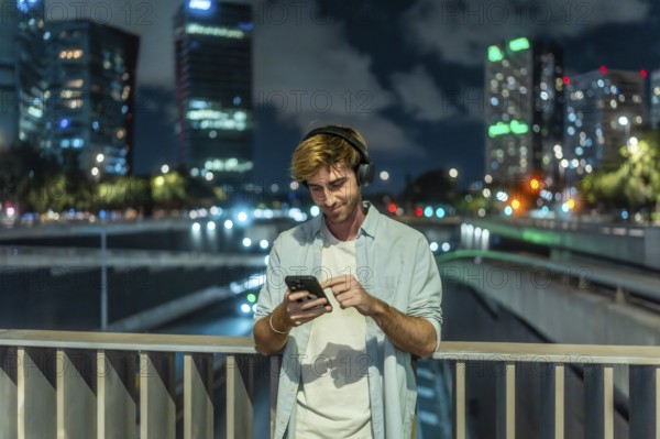 Young man wearing headphones and casual clothing standing on a city bridge at night, smiling and interacting with his smartphone against an urban skyline background