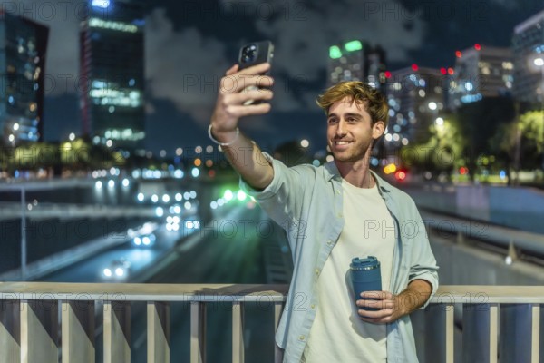 Young man smiling and taking a selfie with a smartphone, holding a reusable coffee cup on an elevated city bridge at night, with traffic lights blurring in the background