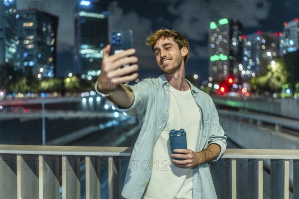 Young man smiling and taking a selfie with a smartphone while holding a reusable coffee cup, standing on a bridge with a glowing city skyline in the background at night