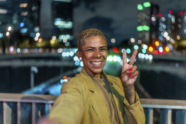 Smiling woman holding up a peace sign, capturing a cheerful selfie moment on a bridge overlooking the illuminated cityscape and vibrant bokeh lights at night