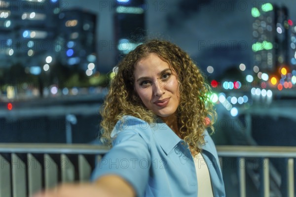 Young woman with curly hair extending her arm to take a selfie, smiling to the camera with blurred city lights and building reflections creating a vibrant bokeh background at night
