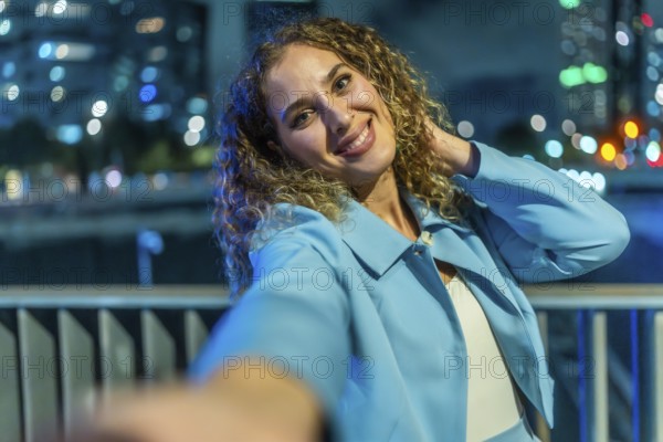 Young woman with curly hair smiling brightly and playfully taking a selfie at night, with a blue jacket and blurred city lights forming a colorful bokeh background