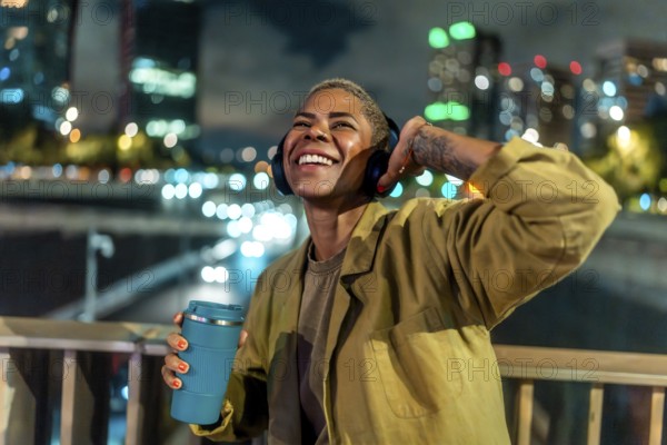 Smiling woman listening to music with headphones, adjusting an earcup, and holding a reusable coffee cup while standing outdoors in a vibrant downtown city at night