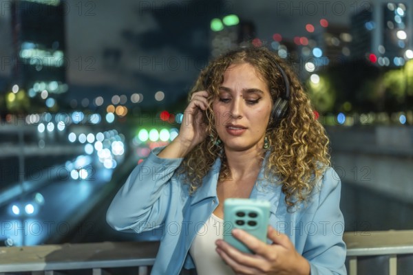 Young woman with curly hair wearing headphones and looking at her smartphone while enjoying music on an urban bridge at night with blurred city lights in the background