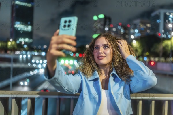 Young woman with curly hair taking a selfie with her mobile phone on a city bridge at night, enjoying urban lifestyle and modern social media communication