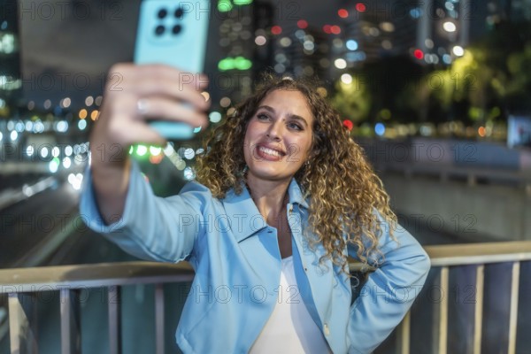 Young woman with curly hair smiling happily while capturing a selfie on her mobile phone, standing on a bridge overlooking city lights and traffic at night