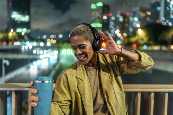 Woman listening to music with headphones and holding a travel mug, smiling brightly while standing on an overpass overlooking a busy city street at night