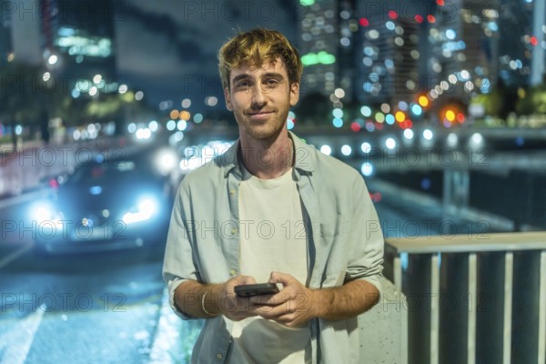 Young man standing outdoors at night smiling at his smartphone, urban downtown bokeh and car lights glowing behind him, relaxed modern lifestyle and digital connectivity vibe
