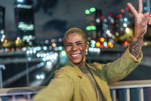Happy young black woman taking a selfie and showing a peace sign, smiling brightly at camera, enjoying a vibrant urban night with blurred city lights in the background