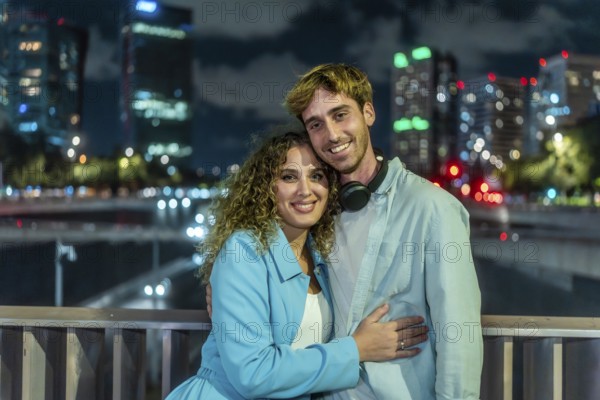 Young couple embracing and smiling at the camera outdoors on a bridge, with illuminated city buildings and bokeh lights creating a romantic urban atmosphere at night