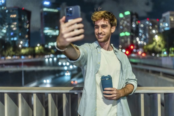 Young man standing on a bridge, holding a reusable cup and smiling while taking a selfie with a smartphone, city skyscrapers and car lights blurring in the background at night