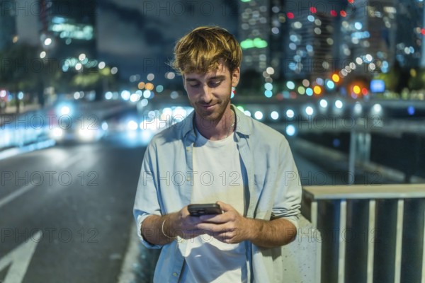Young man standing on a city bridge at night. Engaging with a smartphone. With blurred city lights and moving traffic creating a bokeh effect in the background. Representing modern urban connectivity