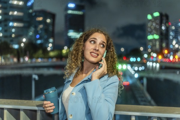 Young woman with curly hair talking on phone with a worried expression while holding a reusable cup, standing on a bridge in an urban city at night with blurred lights