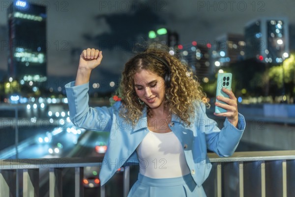 Young woman with curly hair enjoying music through headphones, holding a smartphone, and dancing freely on a bridge overlooking a busy city at night, expressing joy and personal happiness