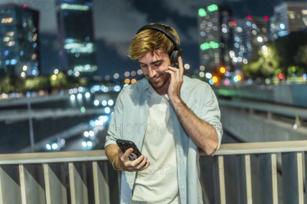 Young man smiling and listening to music through wireless headphones, holding a smartphone while standing on a bridge overlooking a busy city street at night