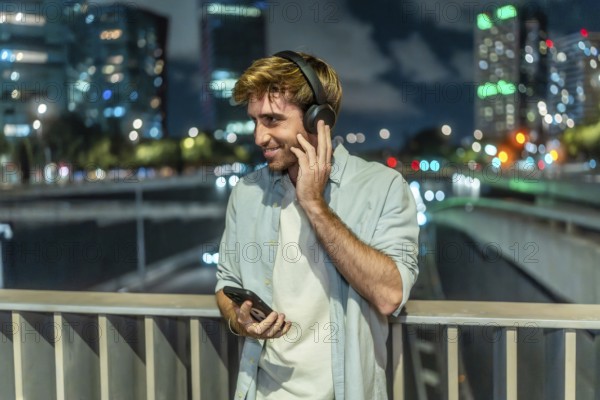 Young man smiling, holding a smartphone and adjusting his headphones on a bridge at night, enjoying personal time with city lights creating a bokeh background