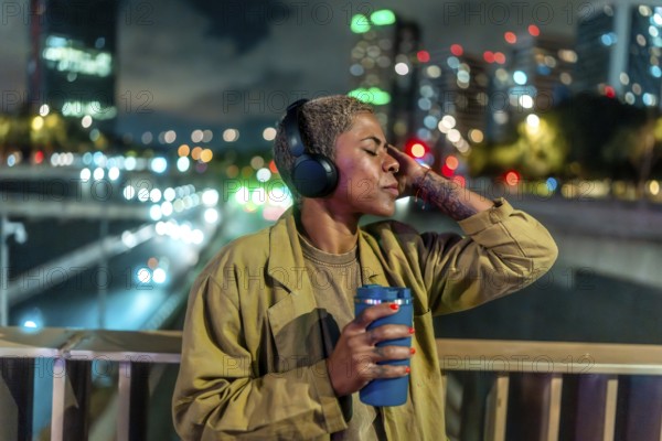 Woman with short hair, wearing headphones and holding a travel mug, relaxing with closed eyes on an urban bridge at night with city lights and bokeh in the background