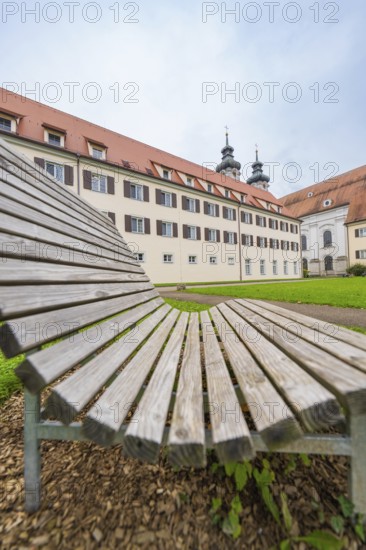 A wooden park bench in the foreground with a monastery building in the background under a cloudy sky, ZFP Zwiefalten, Reutlingen district, Germany