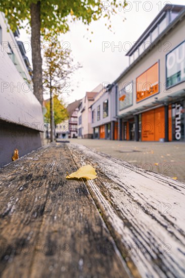 A quiet shopping street with modern buildings, autumn leaves and a wooden bench in the foreground, Nagold, Black Forest, Germany