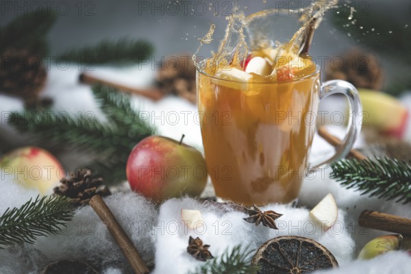 Apple punch splashes out of a glass cup with apple pieces, surrounded by cinnamon sticks and pine branches in a wintry environment
