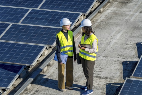 Two diverse female engineers in hard hats and vests discussing an ongoing project while standing on a rooftop surrounded by multiple rows of photovoltaic solar panels