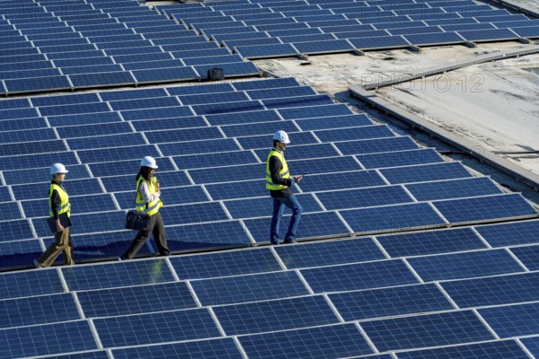Engineers wearing safety helmets and vests are walking across a large array of solar panels, conducting an inspection of the renewable energy installation on a rooftop