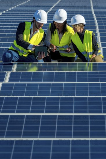 Three diverse engineers in hard hats and vests collaborating while reviewing data on a tablet and clipboard, standing among rows of solar panels at a renewable energy power station