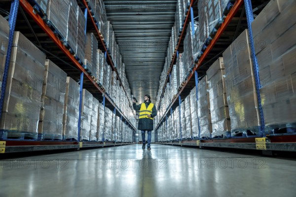 Male worker wearing a safety vest and warm clothing scanning inventory barcodes in a large aisle of a cold storage warehouse, managing frozen logistics and supply chain operations