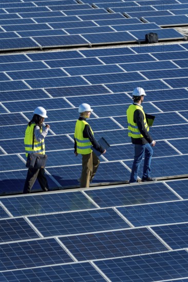Engineers wearing safety vests and hard hats are walking across a vast array of solar panels at a renewable energy power station, conducting inspection and maintenance work