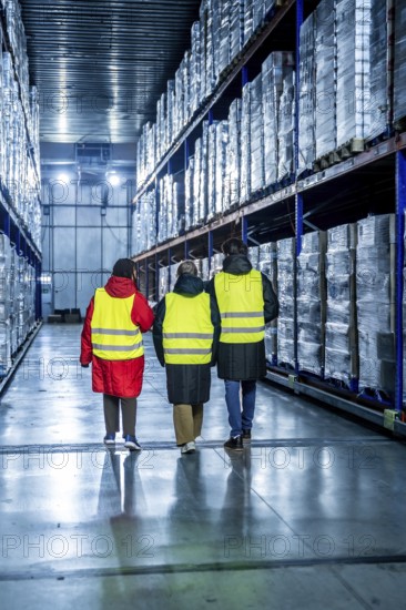 Workers wearing safety vests walking down an aisle in a cold storage warehouse, inspecting pallets of products wrapped in plastic on high racks, managing the supply chain