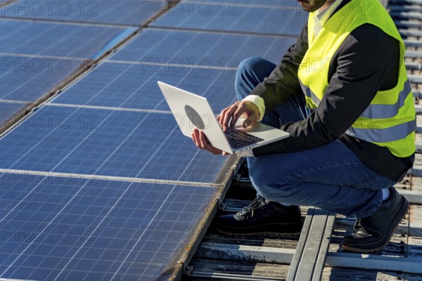 Engineer wearing a high visibility vest and crouching on a rooftop, using a laptop to monitor and check the performance of a large array of solar panels, supporting renewable energy