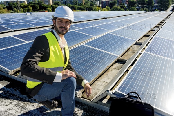 Male engineer in hard hat and safety vest smiling while inspecting rooftop solar panels under blue sky, promoting renewable energy, sustainability and clean power technology