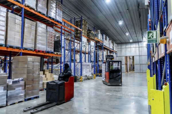Workers are managing inventory with forklifts, navigating between tall shelving units stacked with goods on pallets in a vast, modern cold storage facility, maintaining an efficient supply chain