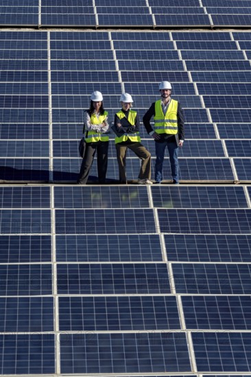 Team of diverse engineers with hard hats and safety vests standing confidently on a large solar panel array, representing renewable energy and sustainable technology