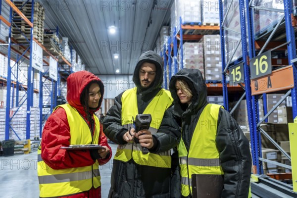 Three warehouse workers in protective clothing and high visibility vests are collaborating while scanning products in a cold storage facility, managing inventory and supply chain operations