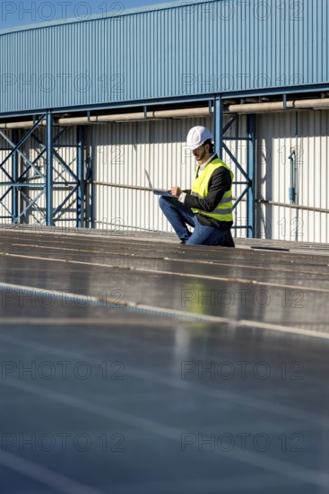 Male engineer wearing a hard hat and safety vest performing an inspection of rooftop solar panels on an industrial building, using a laptop for data management and analysis