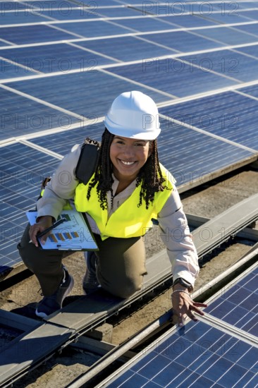 Young woman engineer with safety helmet and vest, smiling while inspecting photovoltaic panels installation on a sunny rooftop, working on sustainable energy and green power