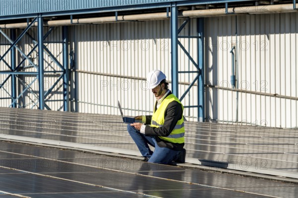 Male engineer inspecting and maintaining solar panel installation on a factory rooftop, capturing data with a laptop to ensure optimal performance and energy efficiency