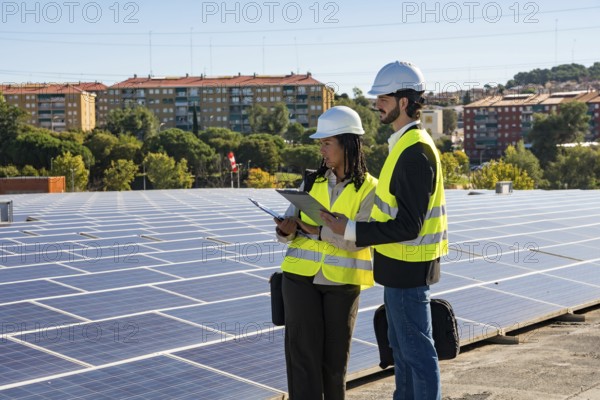 Two engineers wearing hard hats and safety vests reviewing documents on a tablet and clipboard, performing an inspection of a solar panel array on a building rooftop