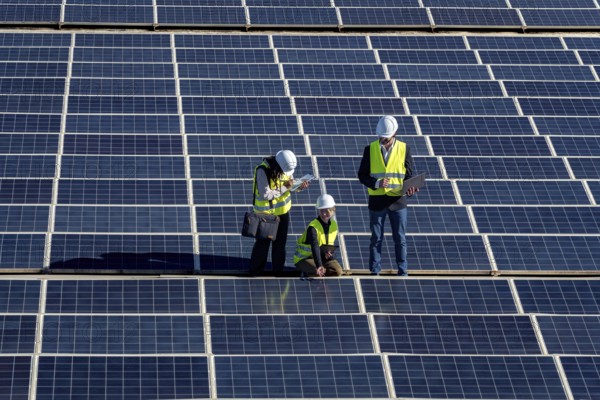 Three engineers inspect a large solar panel array together, checking equipment and performance to maintain efficient photovoltaic production and support renewable energy generation