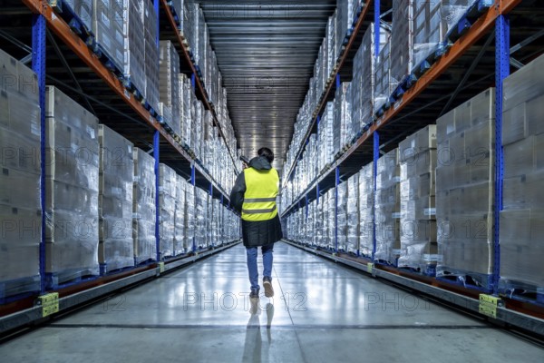 Worker in high visibility clothing walking down a long aisle between rows of elevated racks packed with goods, using a handheld scanner for cold chain logistics and frozen storage management