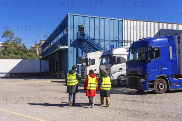 Three workers wearing safety vests walking towards a blue industrial building with multiple semi trucks parked at loading docks, representing cold chain storage and transport operations