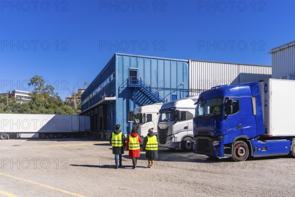 Logistics workers in high visibility vests walking past semi trucks parked at the loading docks of a large industrial warehouse, representing refrigerated transport and cold chain distribution