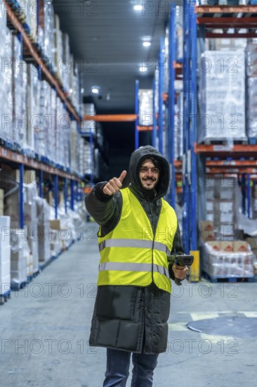 Warehouse worker in warm clothing and safety vest holding a barcode scanner and giving thumbs up inside a cold storage facility, smiling during inventory and logistics operations