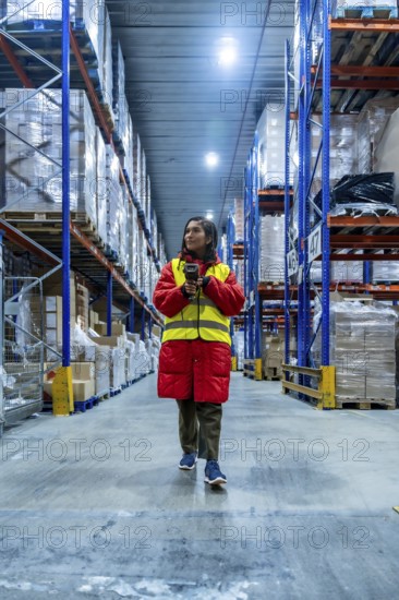 Woman wearing warm jacket and safety vest scanning inventory with a barcode reader, managing stock in a large logistics and distribution cold storage warehouse environment with high racks