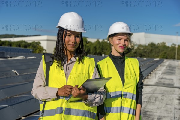 Two women engineers wearing safety vests and hard hats standing on a rooftop with solar panels, focusing on renewable energy and clean power solutions