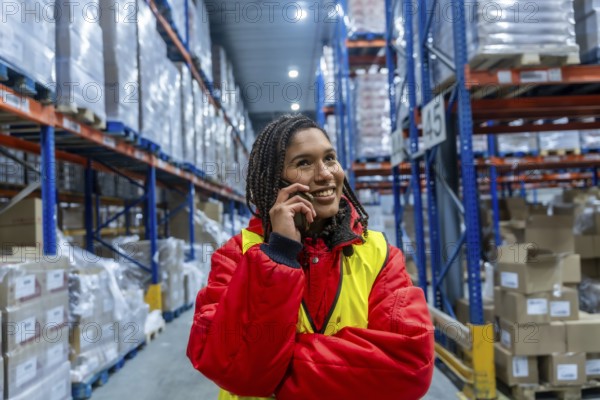 Woman warehouse worker wearing warm clothing and a safety vest, smiling while having a phone conversation inside a large cold storage facility, managing logistics and inventory
