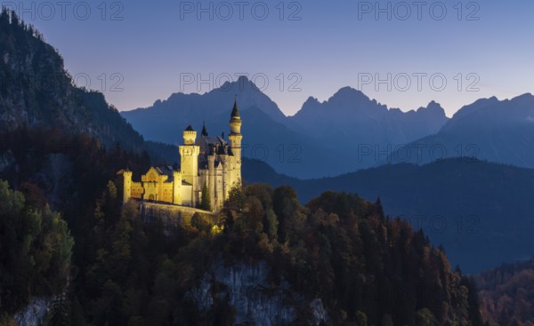 Illuminated Neuschwanstein Castle in front of a rugged mountain panorama, nestled in a dense forest at dusk, Schwangau near Füssen, Ostallgäu, Allgäu, Bavaria, Germany